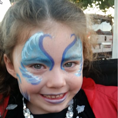 A smiling child with blue, turquoise, purple, and white butterfly-inspired face paint around the eyes, forehead, and cheeks.
