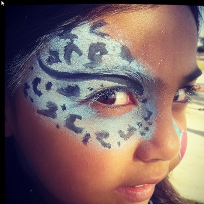A child with a blue-and-black snow-leopard design painted around one eye, finished with glitter.