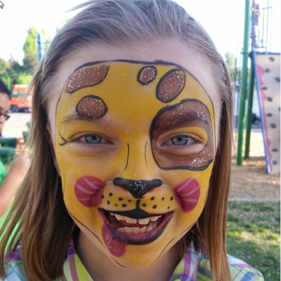 A child at a park wearing a full-face leopard design painted in gold and brown with glitter accents.