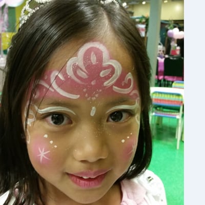 A young girl with pink-and-white floral face paint, white swirls, dots, and glitter accents at an indoor event.