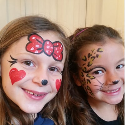 Two smiling children showing colorful bow and animal-themed face paint at an indoor event.